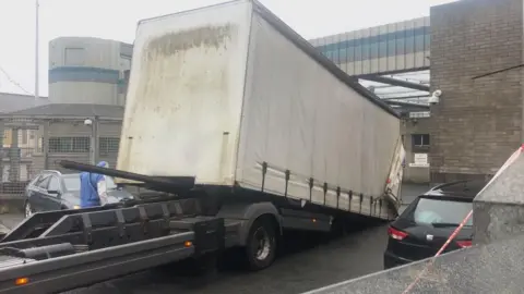 A large white articulated lorry can be seen outside a police station. One person wearing a forensic suit is visible as are two cars