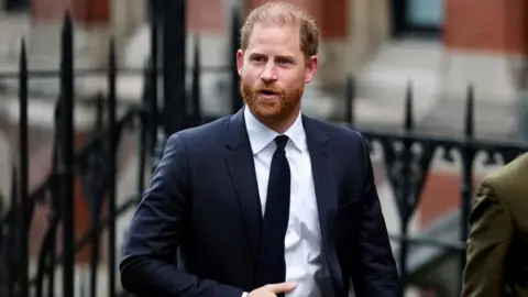 Prince Harry, who has ginger hair and a beard, wears a dark suit and tie with a white shirt as he is seen walking into the High Court building in London