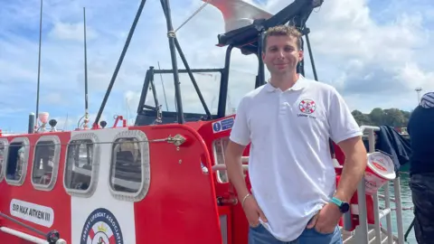 A white man in a Jersey Lifeboat Association t shirt stands in front of a red and white lifeboat called Sir Max Aitken III. 