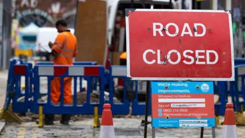 A road closed sign stands in front of utility roadworks on a narrow street, with barriers, traffic cones and workers visible in the background.