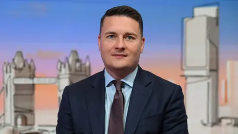 Wes Streeting looks at the camera in a suit and tie from a television set