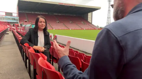 James Vincent / BBC Sports, le ministre Steph Peacock, se trouve avec la BBC Yorkshires James Vincent au Barnsley's Oakwell Stadium. Elle porte une chemise blanche et une veste noire - James Vincent est au premier plan avec une chemise bleue. Ils sont entourés de sièges rouges.