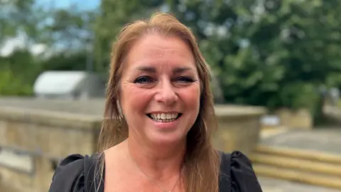 Lisa has long brown hair tied away from her face. She is smiling broadly. She is wearing a black dress and is pictured outside Durham council HQ.