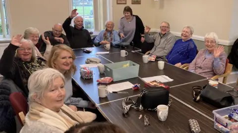 BBC / Simon Spark The photo shows a group of elderly people sitting around a table. They are all smiling at the camera. Some have their arms up, waving.