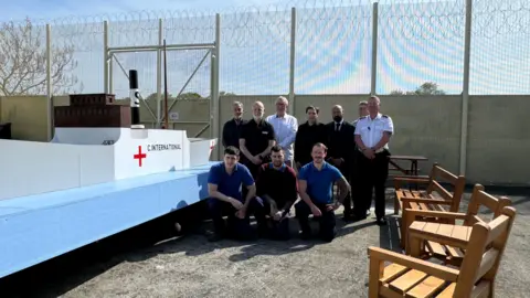 A model of a boat, the SS Vega. It is on a light blue board. The ship is white, with the words C.International on it and a red cross. To the right of the boat are prisoners, kneeling and prison guards stood up and a man in a suit. To the right of them are three benches, faces towards the boat. Behind them is a prison gate.