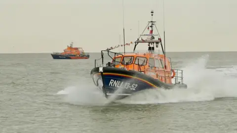 A new lifeboat is speeding through the water at Clacton-on-Sea. It is orange and blue and bears the name RNLI 13-52 painted in white on the side. Sea spray can be seen as it sails through the water