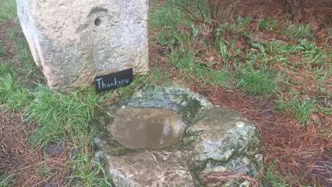 A basin-shaped stone, and filled with brown coloured water, laid flat on the grass. A larger stone is positioned upright above it.