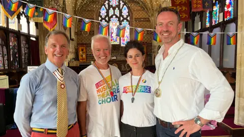 Four people smiling widely inside a church with Pride banners behind them.