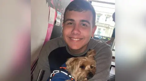 A teenage boy smiles at the camera while a Yorkshire Terrier dog is cuddled up into his arms.