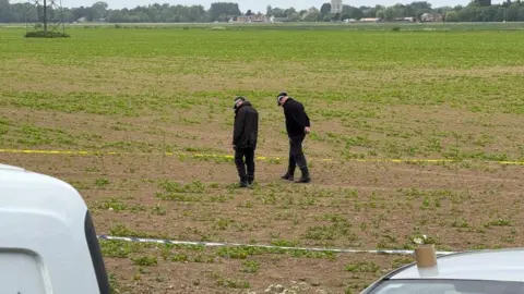 Two police officers walking in a field and peering down at the ground. The area they are in has been taped off with police tape. In the distance, are some buildings and a church steeple.