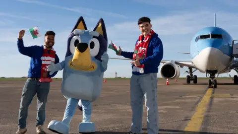 S4C Bluey standing beside two men. They are wearing jeans, blue hoodies and Wales scarves and are holding Welsh flags. All three stand in front of the aeroplane.