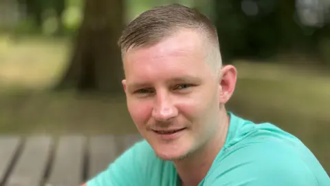 Jack Bennett sonríe para la cámara al aire libre, vistiendo una camiseta de color verde azulado.