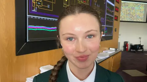 Lauren with brown hair in a plait. She is wearing a green blazer, green tie and white shirt. She is in a classroom. 