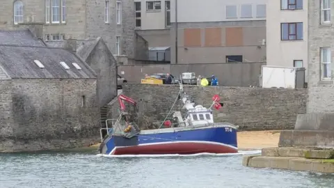 A small red and blue fishing boat aground on a small beach in Lerwick. The sandy beach is surrounded by grey stone and concrete buildings. There are people in the distance overlooking the incident.