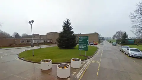 Main entrance to Longcroft School - a grey brick building with a circular grass island at the front with cars parked on one side 
