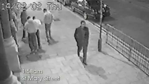 South Wales Police Black and white CCTV image of a man walking down a street - he is clearly seen wearing black in the middle of the screen. A group of other men are to the left. Captions give an indication of where and when the photograph is taken from - Thursday 14/2/02 at 1.58 am on St Mary Street