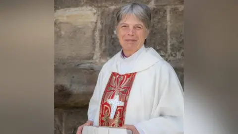 Church in Wales A woman wearing a priest's garb, which is white with a big cross on the front