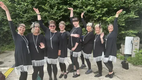 Move to Nurture - Maryam Pourian Eight dance group members stood outside, in front of a hedge, wearing black and silver dance costumes with fringe and a feather headband.
