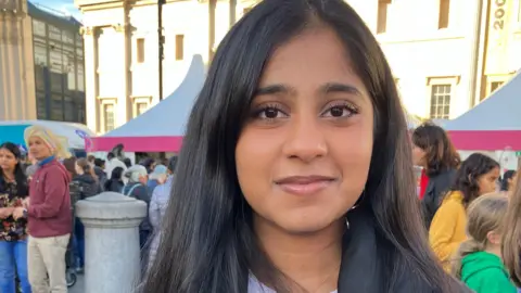 Romilly, a young girl with long dark hair and brown eyes, stood in front of the crowd