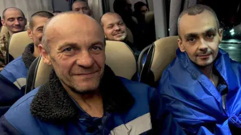 Two newly-released male prisoners of war, in blue jackets, smile at a camera while sat on a coach