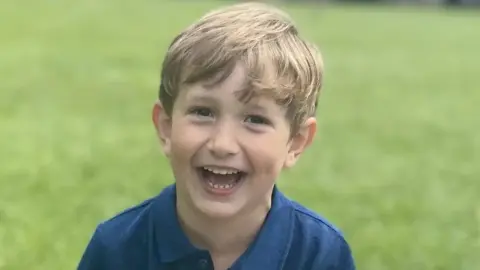 FAMILY HANDOUT Benedict, a young boy with blond hair, is on a lawn. He is wearing a blue polo shirt and is laughing into the camera.