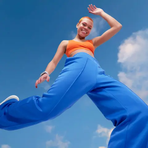 Getty Images Young woman looking energetic and vibrant. She has blue tracksuit bottoms and orange crop top and orange hair. She is striding energetically in front of a blue sky