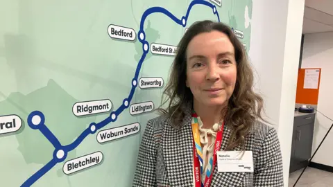 Amy Holmes/BBC A woman stands indoors beside a large wall map showing rail routes and stations, wearing a patterned jacket and an event lanyard. The setting appears to be an exhibition or information space related to transport or infrastructure.