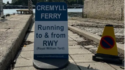 A Cremyll Ferry sign which says the service is running to and from The Royal William Yard. There slipway is in the background and there is a yellow warning cone stopping vehicles from going down to the water.