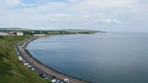 A sweeping bay, viewed from a height. A road along the bay with some caravans and other vehicles parked on it. Steep grass verges beside the road.