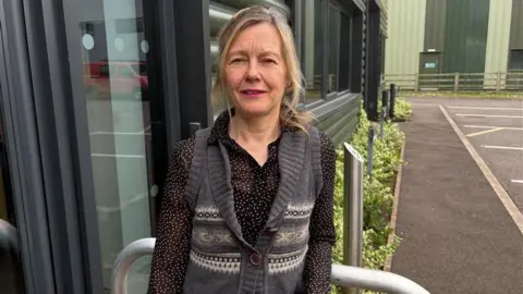 A woman with blonde hair tied back stands in front of a glass door. She is wearing a black blouse with a grey knitted waistcoat. There are shrubs, car parking spaces and a corrugated metal building behind her.
