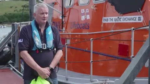 BBC Simon Linsley stood on the left next to a large orange lifeboat. He is wearing a blue float around his neck, a blue RNLI t-shirt and is holding a yellow item.