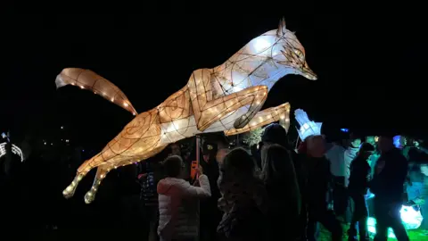 A large, glowing lantern looks like a fox mid-jump with its orange tail in the air. Someone in a white coat is holding the lantern in the air. There are crowds of people in the dark also holding other lit lanterns.