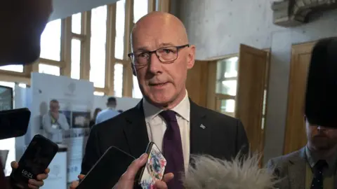 PA Media John Swinney stands in an open area of the Scottish Parliament building while journalists hold their phones and recording devices up to him. He wears glasses and a dark suit
