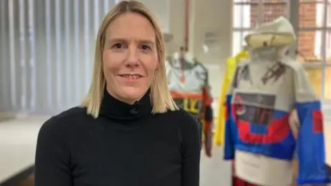 Alex Dunlop/BBC Sarah Graham has blonde shoulder-length hair and wears a black turtleneck top. Behind her are mannequins dressed in colourful garments made from boat sails featuring bold blocks of red, blue, yellow, and white.