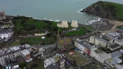 The picture is an aerial photo of a coastal town, showing a mix of buildings, green spaces and cliffs beside the sea. In the foreground there is a cluster of tightly packed houses and shops with pale and pastel‑coloured walls. A tall church with a pointed spire and a roof marked with green and brown patches sits near the centre of the image, surrounded by narrow streets and parked cars.