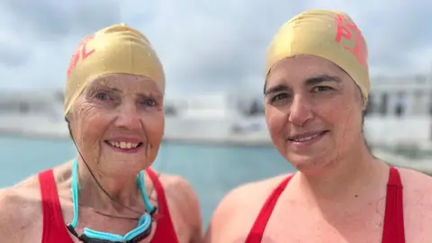 Lesley and Tamsyn Nowman smile at the camera as they stand in front of Penzance's Jubilee Pool, which is blurred behind them. They are wearing gold swimming caps and red swimming costumes. Lesley, who standing on the left hand side, has blue swimming goggles around her neck.