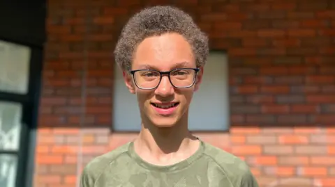 A boy with curly brown hair is wearing glasses and a green camouflage print top. He is standing in front of a brick wall.