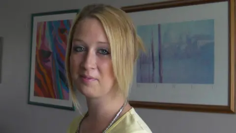 Jasmine Yates A young woman with blonde hair wearing a yellow top in front of two framed art prints.