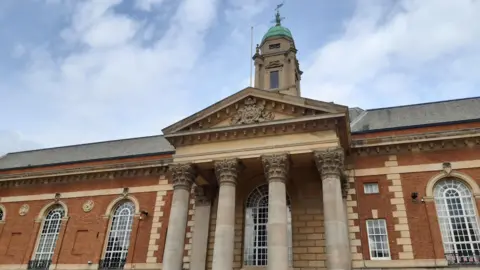 The top of Peterborough town hall. The four pillars outside the entrance are visible. There are blue skies and white whispy clouds.