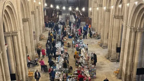 Peterborough Cathedral A picture of the market taken from above inside the Cathedral - with people lining up to buy and view items along white desks placed in lines.