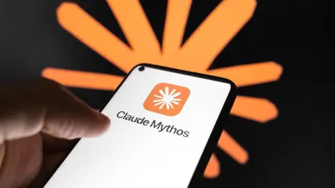 A person's hand holding a smartphone using Anthropic's Claude Mythos app. The words 'Claude Mythos' appear prominently on the white screen with the company logo (a white, cartoonish asterisk on an orange square with curved edges) above. Behind the hand and the phone in soft focus is another version of the logo (an orange, cartoonish asterisk) on a black wall.
