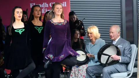 Reuters Three Irish dancers can be seen on stage. Two wearing a black Irish dancing dress and another in purple. They are seen performing for King Charles and Queen Camilla who are picture on the right of them playing bodhran drums. 