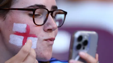 Reuters, a woman wearing glasses wears the England and White flag on her right cheek in the face paint while looking at her reflection on her mobile phone. 