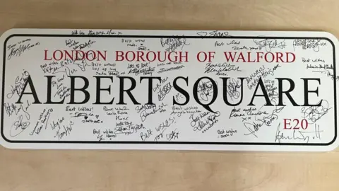 Supplied Shows a sign which reads 'London Borough of Walford, Albert Square, E20'. It's covered in autographs and notes from the cast of EastEnders.