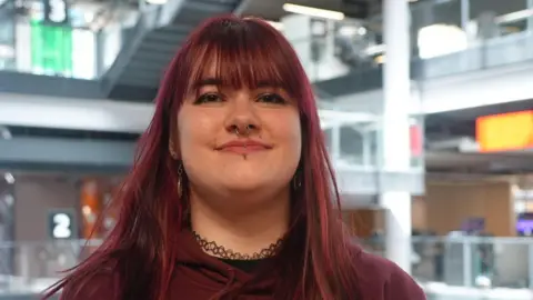 Kady is smiling at the camera. She's wearing a burgundy hoodie and has dyed red hair which is down to her shoulders. She has a nose ring and lip piercings.