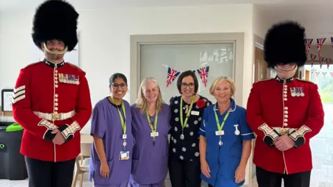 Thames Hospice Two very tall Welsh Guards in red coats and black bearskin hats standing either side of four women wearing medical uniforms. All are smiling and posing for the camera.