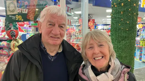 An older man and woman stand in front of a shop inside in shopping centre. The man is wearing a coat and navy jumper over a navy shirt. He has white hair. The woman is wearing a grey scarf with a pink coat. She has grey-blonde hair.