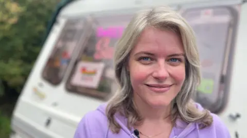 Woman with shoulder-length hair in a mauve top in front of a caravan