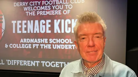 Mickey Bradley is wearing a blue jacket and chequered shirt.
He is standing in front of a large cinema screen that reads: Derry City Football Club welcomes you to the premiere Teenage Kicks - Ardnashee School & College FT The Undertones'.
