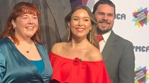 YMCA Exeter A group of people smartly dressed stand on a stage. Two women wearing dresses stand next to each other and smile. Three men dressed in suits are stood behind them. A YMCA Youth Matters Awards backdrop is behind the group.